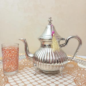 Peut inclure: Une théière argentée avec un pompon décoratif et un verre avec un motif orange sont posés sur un plateau doré. La théière a un bec et une anse incurvés. Le verre est transparent avec un motif complexe.