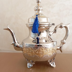 May include: A silver teapot with gold accents and a blue tassel. The teapot has a round body with a decorative design and three legs.