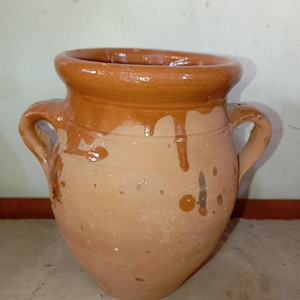 May include: A terracotta pot with two handles. The pot has a wide opening and a rounded body. The top rim and drips are a darker brown than the rest of the pot. The pot is unglazed and has a rustic appearance.