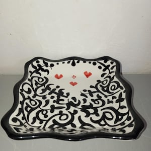 Peut inclure: Un plat décoratif en céramique avec un bord festonné. Le plat présente une base blanche avec un motif floral noir tourbillonnant. Le bord est noir et le centre comporte trois cœurs rouges.