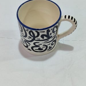Op de afbeelding: Witte keramische mok met een blauwe rand en een zwart abstract ontwerp. De mok heeft een handvat.