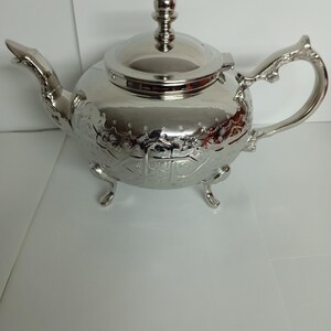 Peut inclure: Une théière en argent avec un design orné et trois pieds. La théière a un couvercle avec un bouton sur le dessus.