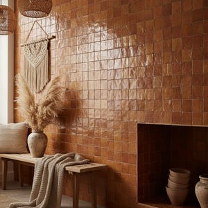 Könnte beinhalten: Ein warmer Innenraum mit einer Wand aus glänzenden, braunen quadratischen Fliesen. Eine Holzbank trägt eine Vase mit getrockneten Pflanzen, ein Kissen und eine Strickdecke. Geflochtene Pendelleuchten und ein Makramee-Wandbehang ergänzen das Bild.