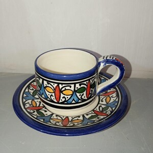 Peut inclure: Un ensemble tasse et soucoupe à café en céramique. La tasse et la soucoupe sont blanches avec un bord bleu et décorées de motifs géométriques colorés en rouge, jaune et vert. La anse de la tasse est bleue avec des accents rouges.
