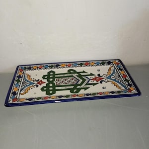 Peut inclure: Plateau de service rectangulaire en céramique avec une base blanche et un motif coloré et complexe. Le plateau présente un motif géométrique vert central, entouré d'une bordure d'éléments décoratifs bleus, oranges, jaunes et verts. Les bords sont bordés d'une bordure bleu foncé.