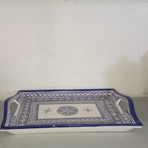 Peut inclure: Plateau de service rectangulaire en céramique avec une base blanche et des motifs géométriques bleus. Le plateau a une bordure et des poignées bleues, avec un motif central de formes stylisées. Pour servir des aliments ou exposer des objets.