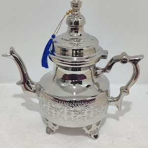 Peut inclure: Une théière en argent avec un design orné et un pompon bleu. La théière a un corps rond avec un bec verseur et une poignée. Elle repose sur trois pieds.