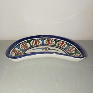 Peut inclure: Un plat en céramique en forme de croissant, avec un intérieur blanc et un bord bleu. Le bord est orné d'un motif répétitif de formes rouges, jaunes, vertes et bleues. Le plat est probablement utilisé pour servir ou exposer.