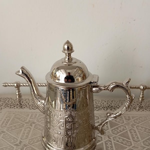Peut inclure: Une théière argentée avec des motifs gravés complexes et une poignée décorative. La théière a un couvercle avec un petit fleuron et un bec verseur incurvé. Elle repose sur un plateau argenté avec un motif géométrique, de style vintage ou antique.
