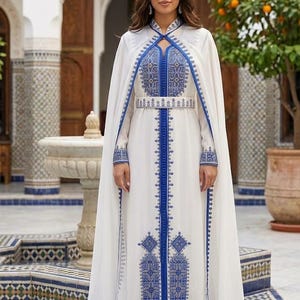 May include: A white dress with blue embroidery and a matching cape. The dress features intricate patterns along the neckline, sleeves, and hem. A silver headpiece adds a touch of elegance. The outfit is suitable for special occasions.
