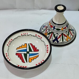 Puede incluir: Una tagine de cerámica blanca con un borde negro y diseños geométricos coloridos. La tapa tiene una forma cónica y la base es un plato poco profundo.