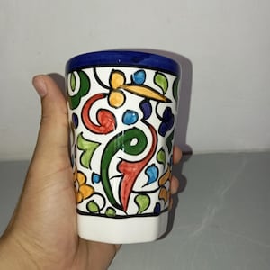 Peut inclure: Une tasse en céramique peinte à la main, avec une base blanche et un bord bleu. La tasse est décorée de motifs floraux et abstraits colorés en rouge, vert, jaune et bleu. La tasse est tenue dans une main.