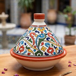 Peut inclure: Un tajine en céramique avec un couvercle conique orné de motifs floraux et de volutes peints à la main en rouge, bleu, vert et jaune sur fond blanc. La base est de couleur crème unie avec un bord rouge et le couvercle a un bouton rouge.