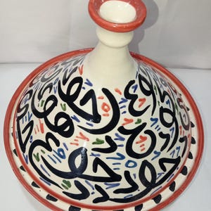 May include: A white ceramic tagine lid with a red rim and black, blue, green, and red Arabic calligraphy design.