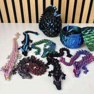 May include: A collection of colorful, articulated dragon figurines and decorative objects. The dragons are in various colors, including pink, blue, green, and purple. One dragon is coiled, and another is inside a blue and green container. The background is a white surface.