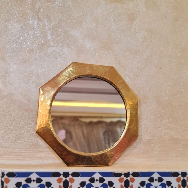 May include: A small octagonal mirror with a hammered gold finish. The mirror is surrounded by a decorative frame and reflects a room with a white ceiling and a patterned curtain.