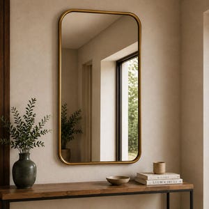 May include: A rectangular wall mirror with rounded corners and a thin gold-toned metal frame. The mirror is mounted on a light-coloured wall above a wooden console table that holds a dark vase with greenery, a small bowl, and two stacked books.