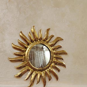 K&ouml;nnte beinhalten: Ein goldener Sonnenspiegel mit einem runden Spiegel in der Mitte. Der Spiegel ist von einem dekorativen Sonnenstrahlendesign umgeben.