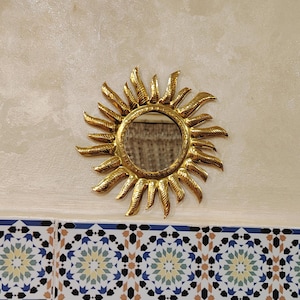 K&ouml;nnte beinhalten: Ein goldener Sonnenspiegel mit einem runden Spiegel in der Mitte. Der Spiegel h&auml;ngt an einer Wand mit wei&szlig;en und beigen Fliesen.