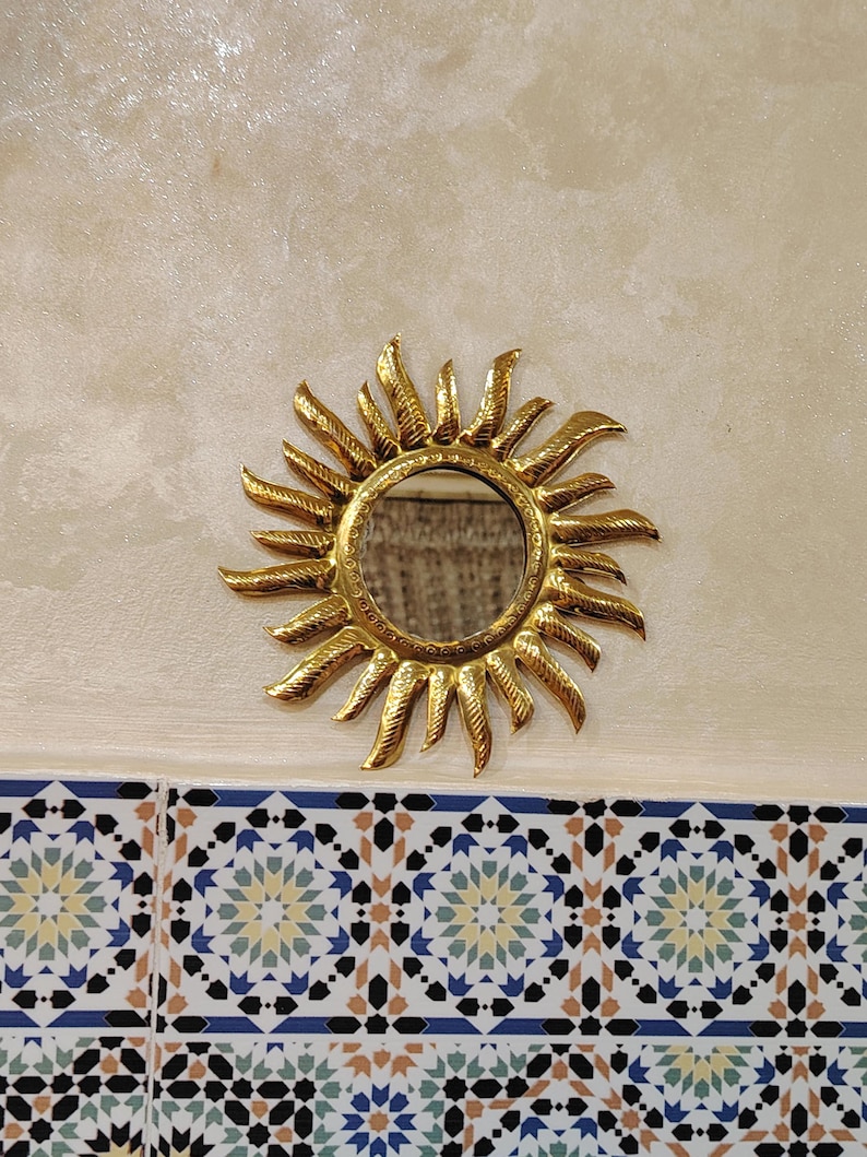 K&ouml;nnte beinhalten: Ein goldener Sonnenspiegel mit einem runden Spiegel in der Mitte. Der Spiegel h&auml;ngt an einer Wand mit wei&szlig;en und beigen Fliesen.
