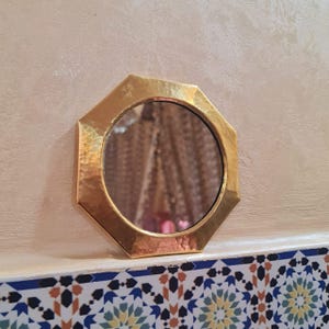 May include: A small, octagonal, gold-toned mirror with a hammered finish. The mirror is reflecting a blurry image of a room with a patterned rug.