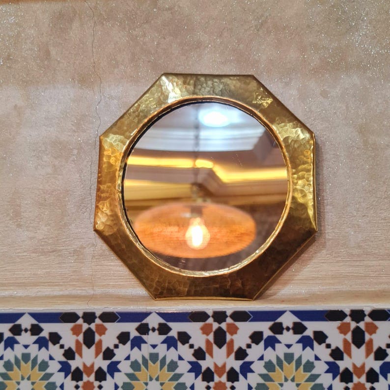 May include: A gold octagonal mirror with a hammered finish. The mirror reflects a light fixture with a light bulb.