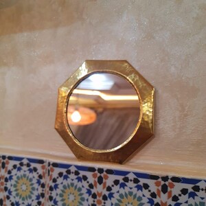 May include: A small, octagonal, gold-toned mirror with a hammered finish. The mirror is mounted on a wall with a tiled surface below.