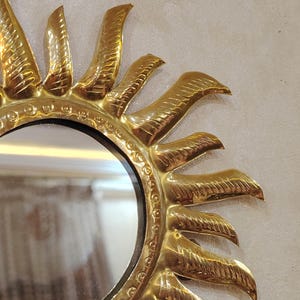 K&ouml;nnte beinhalten: Ein goldfarbener Sonnenspiegel mit einem runden Spiegel in der Mitte. Der Spiegel hat einen dekorativen Rand aus kleinen Kreisen und die Sonnenstrahlen haben ein strukturiertes Design.