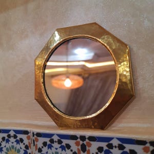 May include: Octagonal brass mirror with a hammered finish. The mirror has a slightly rounded edge and reflects a room with a light fixture.