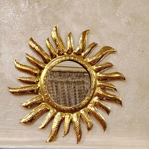 K&ouml;nnte beinhalten: Ein goldener Sonnenspiegel mit einem runden Spiegel in der Mitte. Der Spiegel ist von einem dekorativen Metallrahmen mit einem Sonnenstrahlendesign umgeben.