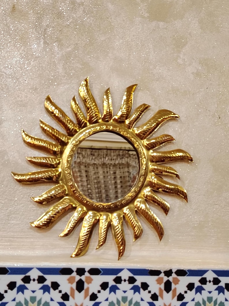 K&ouml;nnte beinhalten: Ein goldener Sonnenspiegel mit einem runden Spiegel in der Mitte. Der Spiegel ist von einem dekorativen Metallrahmen mit einem Sonnenstrahlendesign umgeben.