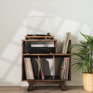 Peut inclure: Un meuble de rangement pour disques vinyles en bois brun foncé avec quatre pieds, comprenant une étagère supérieure pour une platine et un amplificateur, ainsi que trois compartiments inférieurs. Le meuble possède une structure rectangulaire.