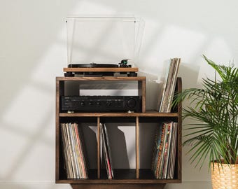 Puede incluir: Un soporte de madera oscura para tocadiscos con una superficie superior para el plato, un estante central con un receptor de audio negro y tres compartimentos verticales inferiores llenos de discos de vinilo, sobre cuatro patas inclinadas.