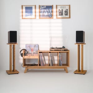 Op de afbeelding: Een mediameubel van licht hout met drie verticale vakken voor vinylplaten, een platenspeler bovenop en twee bijpassende luidsprekerstandaards met zwarte luidsprekers. Drie ingelijste vinylhoezen hangen aan de muur boven het meubel.