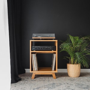Peut inclure: Un meuble multimédia en bois clair à trois niveaux. Le niveau supérieur accueille une platine vinyle noire, le niveau intermédiaire contient un amplificateur noir et le niveau inférieur range des disques vinyles à la verticale.