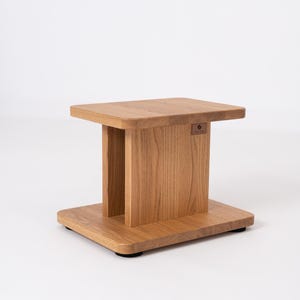 Può includere: Uno stand per altoparlanti in legno marrone chiaro con una piattaforma superiore rettangolare e una base abbinata collegate da un pannello di supporto verticale centrale. Una piccola etichetta rettangolare del marchio è fissata sul lato e sono visibili piedini circolari neri.