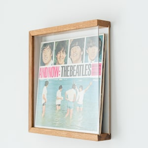 May include: A square wooden frame holding a vinyl record cover titled And Now: The Beatles. The cover features four headshots of the band members above an image of the group standing in shallow water. The frame has a clear front panel.