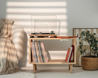 May include: A light-colored wooden record player stand with four short legs, featuring an open shelf filled with vinyl records and a few books. A black turntable with a clear plastic dust cover sits on the top surface of the wooden stand.