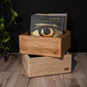 Può includere: Due cassette rettangolari in legno impilate verticalmente. La cassetta superiore è realizzata in rovere scuro e contiene una collezione di dischi in vinile, tra cui uno con il testo COMA Pierwsze wyjście z mroku. La cassetta inferiore è in legno chiaro.