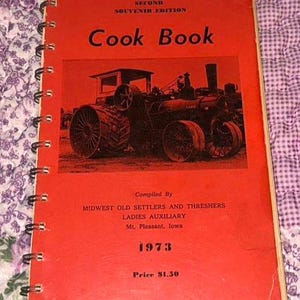 Può includere: Un libro di cucina a spirale rosso intitolato Second Souvenir Edition Cook Book, con un'illustrazione in bianco e nero di un trattore a vapore d'epoca. La copertina include il testo che identifica i compilatori come Midwest Old Settlers and Threshers Ladies Auxiliary, Mt. Pleasant, Iowa, l'anno 1973 e un prezzo di 1,50 $.