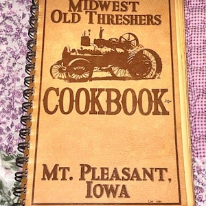 Può includere: Un ricettario con rilegatura a spirale e copertina color marrone chiaro con l'illustrazione di un trattore a vapore d'epoca. Il testo riporta MIDWEST OLD THRESHERS COOKBOOK e MT. PLEASANT, IOWA, con le iniziali LM e l'anno 1980 nell'angolo.