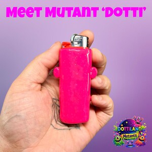 May include: A vibrant pink lighter with a red top and silver metal details. The lighter features two small, rounded side protrusions. The text "Meet Mutant 'Dotti'" is visible above the lighter.