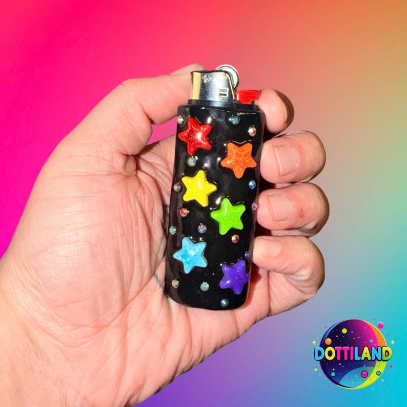 May include: Black lighter decorated with colorful star-shaped embellishments. The stars are red, orange, yellow, green, blue, and purple. The lighter also has small, clear rhinestones. The lighter is held in a hand.