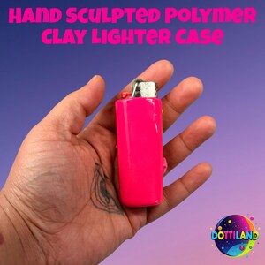 Op de afbeelding: Een felroze polymeerklei aanstekerhoes. De aanstekerhoes wordt in een hand gehouden tegen een paarse en blauwe achtergrond. De tekst "HAND SCULPTED POLYMER CLAY LIGHTER CASE" staat bovenaan de afbeelding. Het Dottiland-logo staat rechtsonder.
