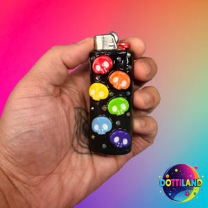 K&ouml;nnte beinhalten: Schwarzes Feuerzeug, verziert mit bunten Totenkopf-Designs. Die Totenk&ouml;pfe sind rot, orange, gelb, gr&uuml;n, blau und lila. Das Feuerzeug hat einen silbernen Metallaufsatz und ist mit kleinen, funkelnden Akzenten verziert.