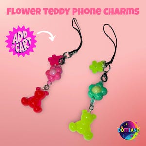 May include: Two phone charms with the text "FLOWER TEDDY PHONE CHARMS". One charm features a red teddy bear and pink flower accents. The other charm has a green teddy bear and green flower accents. Both charms have black straps.