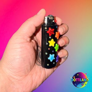 May include: A black lighter decorated with colorful star-shaped embellishments. The stars are red, orange, yellow, green, blue, and purple. Small, iridescent gems are scattered around the stars. The lighter is held in a hand against a gradient background.