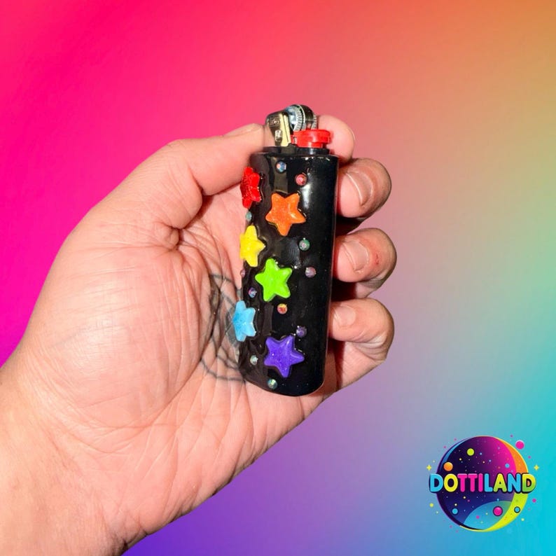 May include: A black lighter decorated with colorful star-shaped embellishments. The stars are red, orange, yellow, green, blue, and purple. The lighter is held in a hand against a gradient background of pink, purple, and blue.