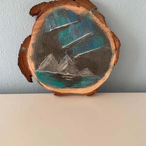 Puede incluir: Una rodaja de madera pintada a mano con una escena de paisaje. La obra de arte representa montañas, agua y un cielo nocturno con vetas de color. La madera tiene un borde natural y rústico.