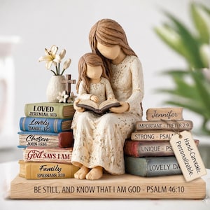 May include: A figurine of a mother and child reading a book, seated on a stack of books with titles like "Loved" and "Family." A vase with flowers and a cross are also present. The base reads "Be Still, and Know That I Am God."